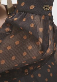 Manche en tissu noir transparent avec des pois marron et un détail de bouton en laiton sur le poignet.