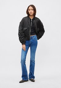 Fekete bombázó dzseki bordázott mandzsettával és szatén hatású anyagból, bővülő kék farmernadrággal és szegecses, hegyes orrú fekete cipővel párosítva.