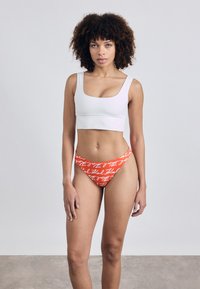 Bílý crop top v kombinaci s oranžovými spodními kalhotkami s bílým vzorem nápisů. Hladká tkanina s přiléhavým střihem a minimálními švy.
