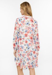 Una donna con lunghi capelli castano chiaro indossa un vestito floreale colorato, vista da dietro su uno sfondo bianco.