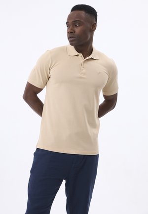 Camisa de polo beige con cuello y tres botones, con un pequeño logo en el lado izquierdo del pecho, combinada con pantalones azul marino. Textura suave.