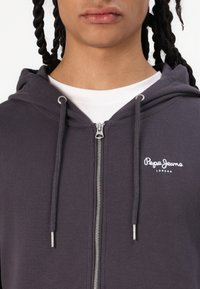 Sudadera con cremallera de color gris oscuro, con una cremallera plateada, que presenta el logo blanco de "Pepe Jeans London" en el pecho y cordones ajustables en la capucha.