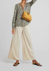 Femme portant un chemisier vert à motifs de feuilles, un pantalon large crème, des sandales noires à lanières, et portant un petit sac à main tressé beige avec une bandoulière.