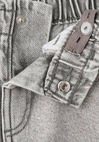 Harmaa farkkukangas, jossa on nappi- ja nepparikiinnitys. Näkyvät saumat ja pehmeä, hieman teksturoitu viimeistely korostavat designia.