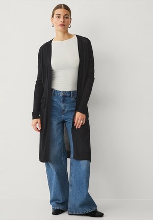 Zwarte lange cardigan met open voorkant, gedragen over een witte top, gecombineerd met hoge taille blauwe wijde jeans en zwarte schoenen. Geglansde stoftextuur.