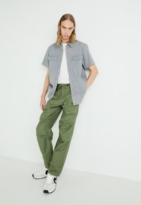 Camisa de manga corta gris claro con botones para hombre, con dos bolsillos en el pecho, combinada con pantalones cargo holgados de color verde olivo y zapatillas blancas.