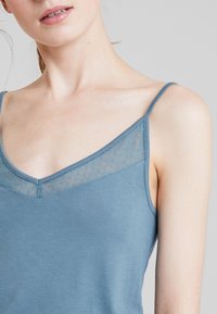 Blå linne med V-krage, med genomskinliga meshpaneler med små prickmönster vid axlarna. Mjuk tyg med justerbara axelband.