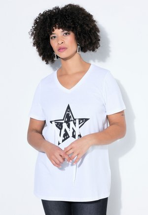 Frau mit lockigem Haar, die ein weißes V-Ausschnitt-T-Shirt mit einem schwarzen Stern und Buchstabenmuster trägt, vor einem einfarbigen weißen Hintergrund.