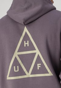 Sudadera morada con un gran gráfico triangular en color crema en la parte posterior, que muestra las letras H, U y F dentro de los triángulos.