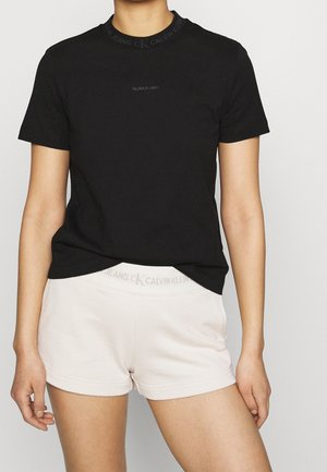 Person, die ein schwarzes Calvin Klein T-Shirt und hellbeige Calvin Klein Shorts vor einem einfarbigen Hintergrund trägt.