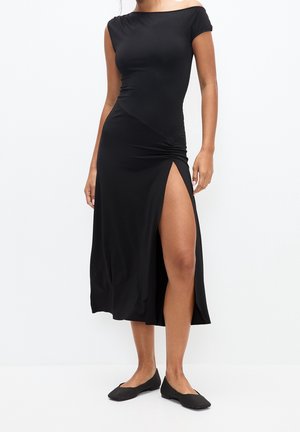 Femme portant une robe noire cintrée, épaules dénudées, avec une fente haute sur la cuisse gauche et des chaussures plates noires, debout devant un fond blanc.