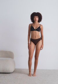 Mustikan pitsirajoitusmustalla lace-bralette ja ruskeat teksturoidut korkeavyötäröiset alushousut mallilla. Pehmeä, neutraali tausta kaarevalla sohva.