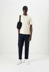 Beige cotton t-shirt, navy trousers, white sneakers, black crossbody bag, black cap; model stands against a white background.