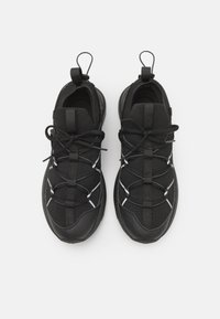 Baskets de sport noires avec un upper en maille texturée, des lacets élastiques et une semelle rembourrée. Des accents réfléchissants ajoutent un détail visuel. Vue de dessus.