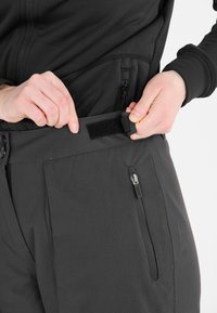 Zwarte outdoorbroek met een gladde textuur, voorzien van een ritszak en een met klittenband verstelbare tailleband. Close-up van de taillebandverstelling.