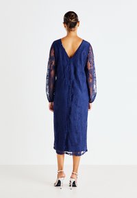 Vestido de renda azul-marinho com mangas longas e transparentes e design em V nas costas. Apresenta padrões florais intrincados e um corte a meio comprimento. Sandálias brancas de salto.