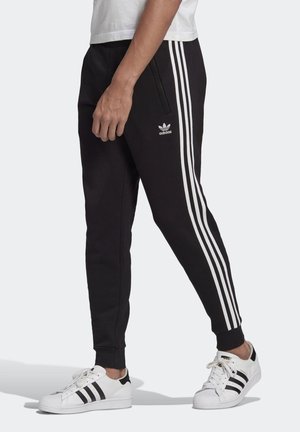 Person trägt schwarze Adidas-Trainingshose mit weißen Streifen und Logo, kombiniert mit weißen Adidas-Sneakern mit schwarzen Streifen.
