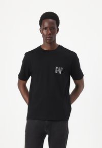 Camiseta de algodón negra con un pequeño logo blanco de "GAP" en el lado izquierdo del pecho, cuello redondo y mangas cortas. Combinada con jeans oscuros.