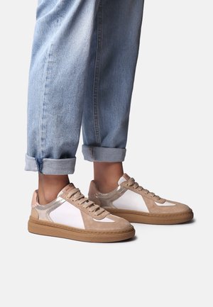 Piedi con indosso sneaker basse beige e bianche abbinate a jeans di denim azzurro chiaro risvoltati su uno sfondo semplice.