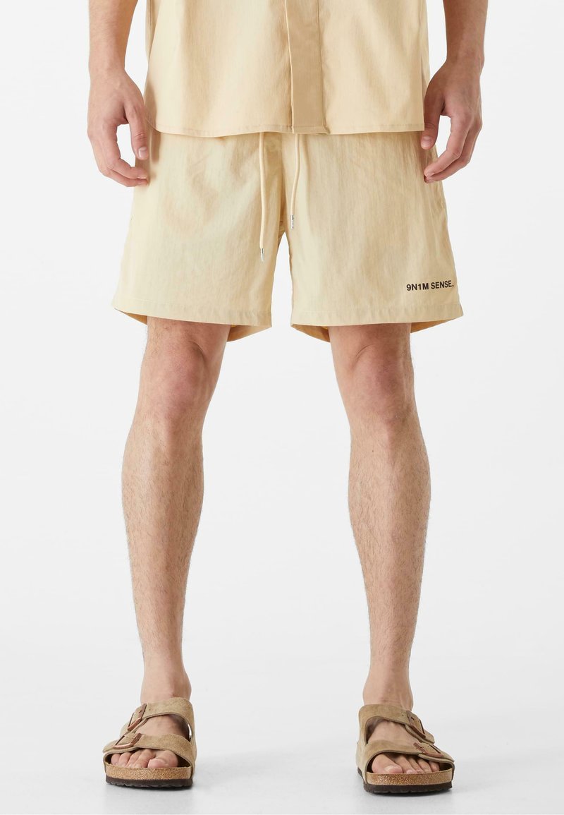 Krátke béžové šortky s pásom na stiahnutie a detailom loga. Nosené s hnedými sandálmi, ktoré predvádzajú hladký povrch a uvoľnený strih.