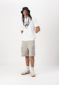 Hvid t-shirt med et sort grafisk design, tan cargo shorts, grå bucket hat og beige sneakers med brune accenter. Enkel baggrund.