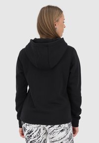 Felpa nera con cappuccio e maniche lunghe, caratterizzata da una texture morbida e una vestibilità comoda. Indossata con leggings bianchi e neri a fantasia.