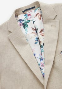 Blazer de lino beige que muestra un forro de estampado floral en colores vibrantes. Presenta un solapa con muesca y un detalle de bolsillo en el pecho.