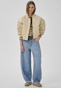 Puffige beige Bomberjacke, kariertes Hemd darunter, hellblaue Jeans mit weitem Bein und braune Slipper mit ausgefransten Säumen.