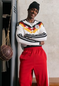 Ung man ler med armarna i kors, b är en svart mössa, Tysklands fotbollströja och röda träningsbyxor, står vid ett öppet skåp.