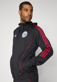 Schwarzer Kapuzenpullover mit roten Akzenten und drei roten Streifen an den Ärmeln. Mit einem Logo auf der Brust und seitlichen Taschen. Glattes, sportliches Material.
