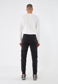 Man som vänder bort, klädd i en vit långärmad skjorta, svarta cargobyxor med sidofickor och grå sneakers mot en enfärgad bakgrund.