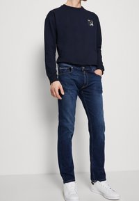 Camisa de manga larga azul marino con un pequeño parche de logo, combinada con unos vaqueros ajustados de color azul oscuro y zapatillas blancas. Diseño casual y sencillo.