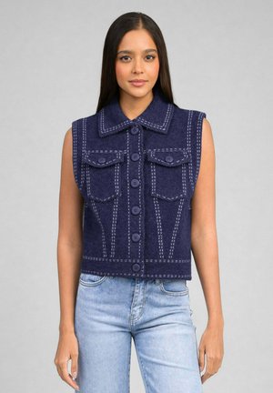 Jeune femme aux longs cheveux foncés porte un gilet sans manches bleu marine boutonné avec des coutures blanches et un jean bleu clair sur fond gris.