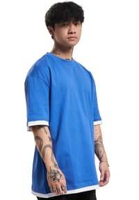 T-shirt en coton bleu avec des poignets et un col blancs. Présente une coupe ample et des manches courtes. Des tatouages remarquables sur le bras sont visibles.
