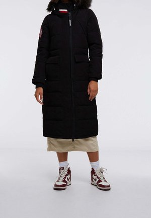 Personne portant un long manteau matelassé noir avec capuche en fourrure, jupe beige, chaussettes blanches et baskets hautes rouges et blanches sur un fond uni.