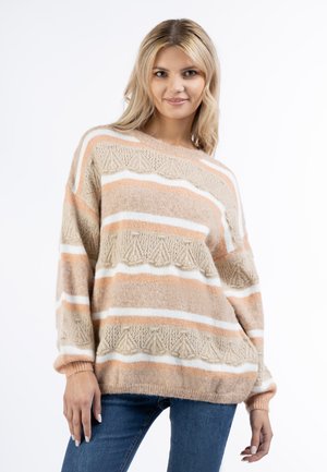 Donna bionda che indossa un maglione a righe beige, bianco e pesca con motivi strutturati, posando con una mano in tasca e jeans blu.