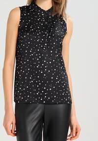 Haut sans manches noir avec un motif d'étoiles argentées. Présente un col à volants et un détail de boutons. Le matériau semble lisse et brillant.