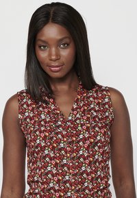 Blusa sin mangas con un estampado floral rojo que presenta flores blancas, rosas y amarillas. La tela es ligera, con un cierre de botones en la parte delantera y un cuello.