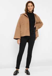 Manteau en laine à capuche marron avec une silhouette carrée et de grandes poches avant, associé à un pantalon noir ajusté et des mocassins noirs.