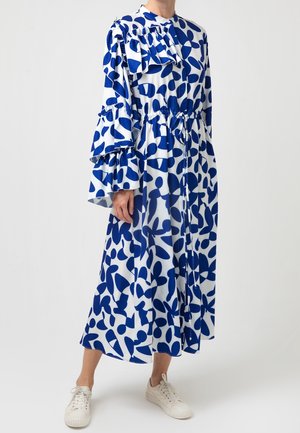 Langes weißes Kleid mit auffälligen blauen abstrakten Formen, gerafftem hohem Kragen, mehrlagigen Glockenärmeln, taillierter Taille mit Bindeband, getragen mit weißen Sneakern.