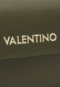 Närbild av strukturerad mörkgrön läder med silvermetallisk Valentino-logotyp och synliga sömdetaljer.