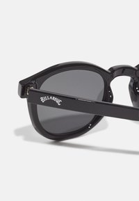 Schwarze Sonnenbrille mit hochglanzmäßiger Oberfläche, runden Gläsern und dem weißen "Billabong"-Logo am Bügel. Dunkelgraue getönte Gläser.