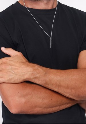 Homme en chemise noire à manches courtes, les bras croisés, portant une chaîne en argent avec un pendentif vertical à motif chevron.
