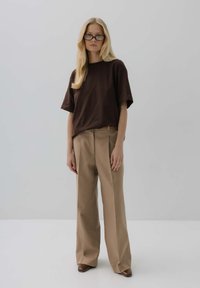 Brązowa, oversize bawełniana koszulka zestawiona z beżowymi spodniami o wysokim stanie, z prostym krojem i kieszeniami oraz brązowymi butami na obcasie.
