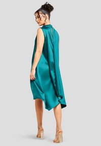 Vestido sin mangas de satén en tono verde azulado con un escote alto, espalda drapeada, dobladillo asimétrico y un detalle de ojal en el cuello. Sandalias de tacón transparente.