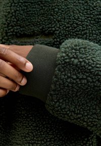 Veste en polaire vert foncé avec un extérieur texturé et duveteux et des poignets côtelés. Vue en gros plan d'une main reposant sur la manche.