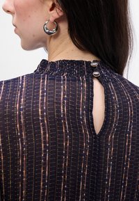 Blusa nera a motivi con strisce verticali in oro, blu e rosa. Presenta un collo alto smock e un'apertura a chiave sul retro con bottoni.