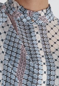 Blusa trasparente con colletto a ruffles, motivi geometrici nei colori blu, rosa e bianco, con dettagli metallici e chiusura a bottoni.