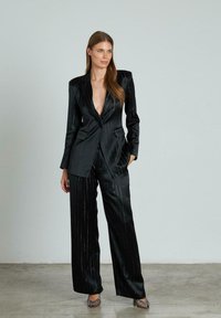 Completo a righe verticali nere realizzato in tessuto liscio, con blazer a profondo scollo a V e pantaloni a gamba larga. Accentuato da strisce verticali lucide.