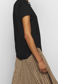 Musta lyhythihainen paita pyöreällä kaula-aukolla, yhdistettynä ruskeaan pukuhameeseen, jossa on houndstooth-kuvio.